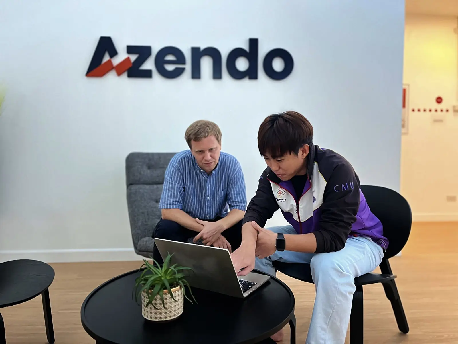 Two offshore developers working on a laptop in front of the Azendo logo wall